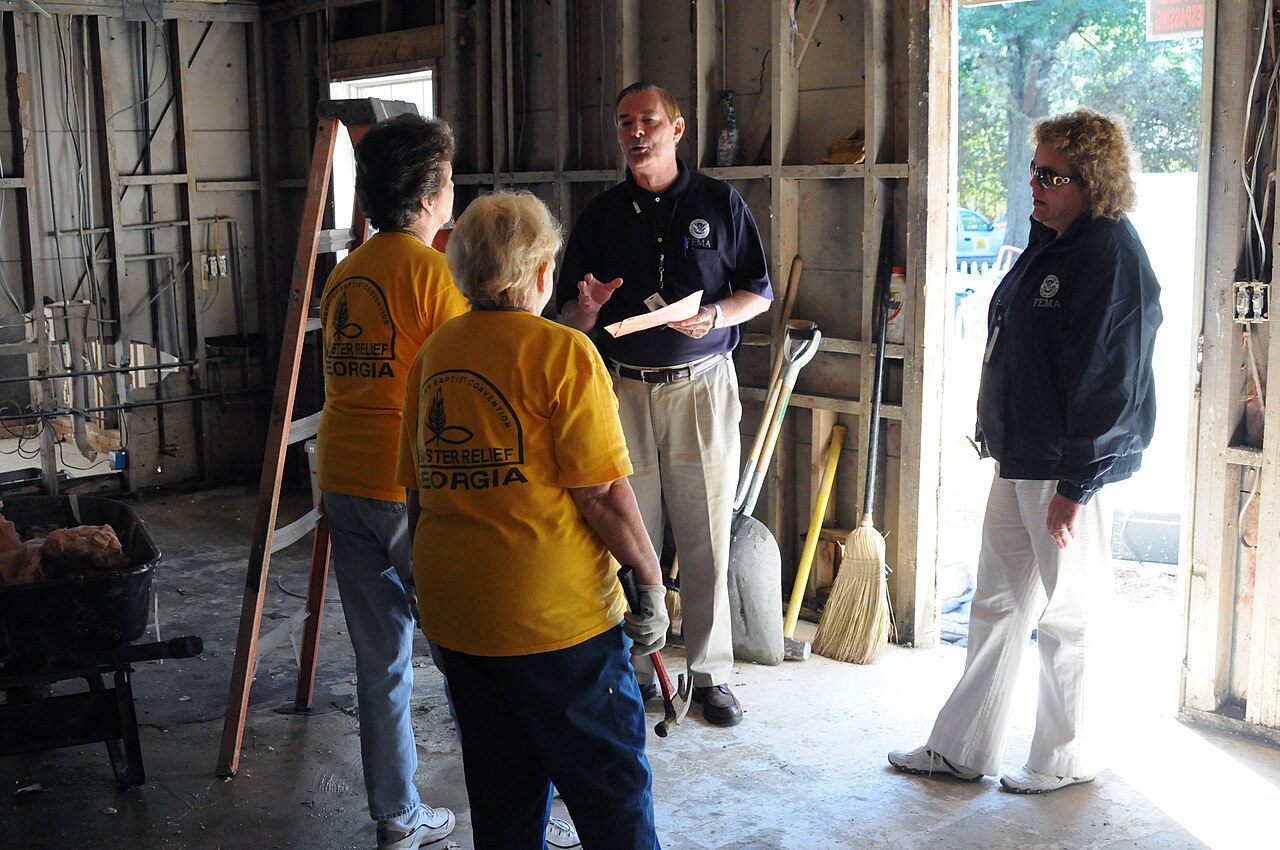 Volunteers in yellow shirts speaking with officials inside a stripped residential room (FEMA photo by Andrea Booher)